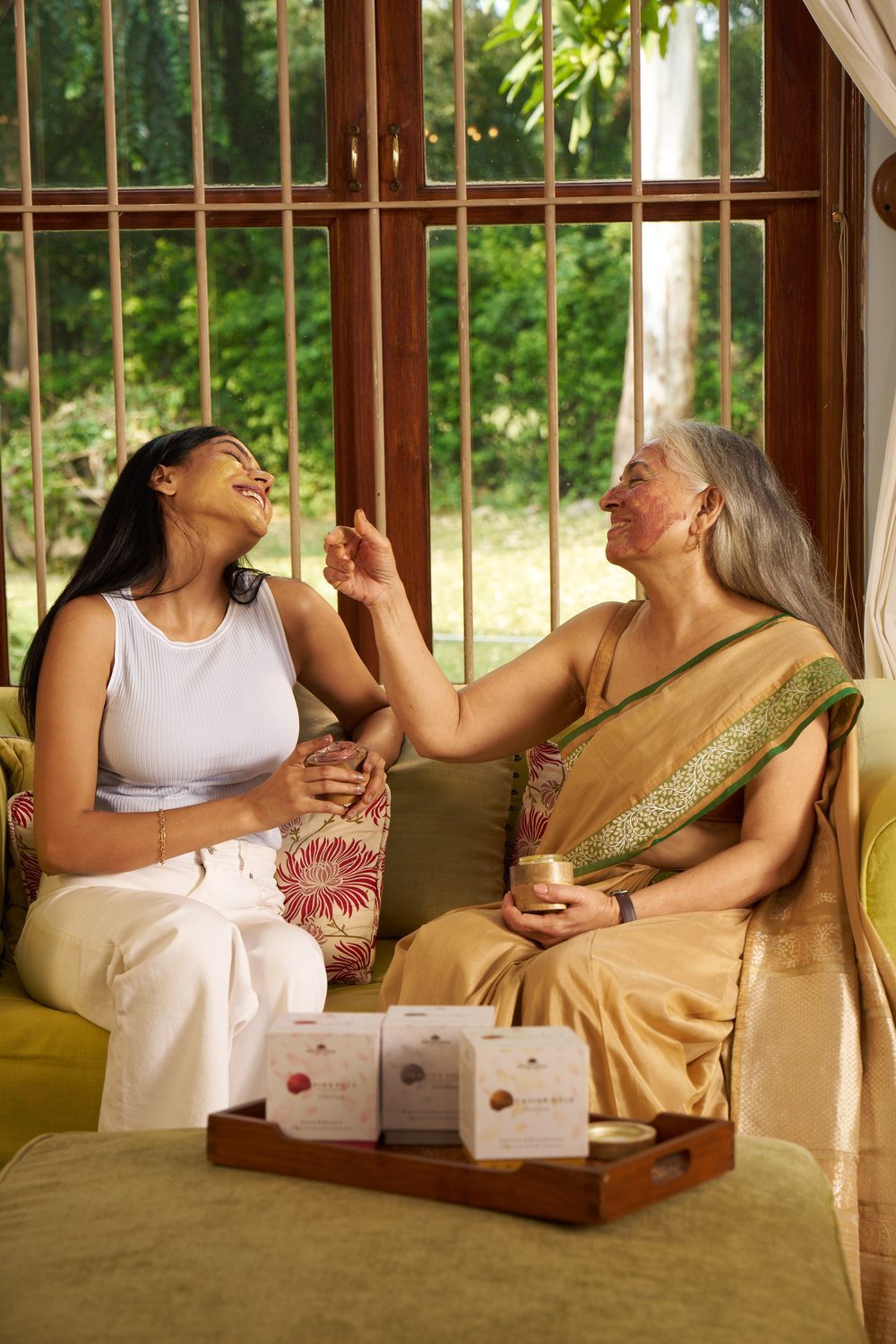 Two women laughing with face packs on, products on a tray in the foreground.